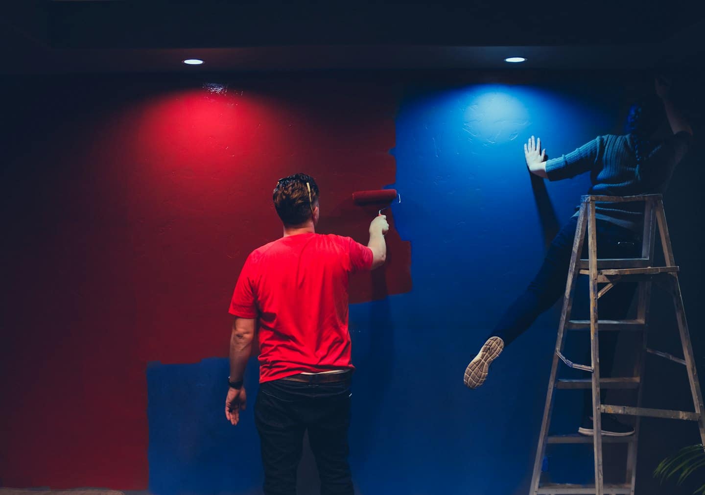 Man paints wall red while person on ladder paints blue.