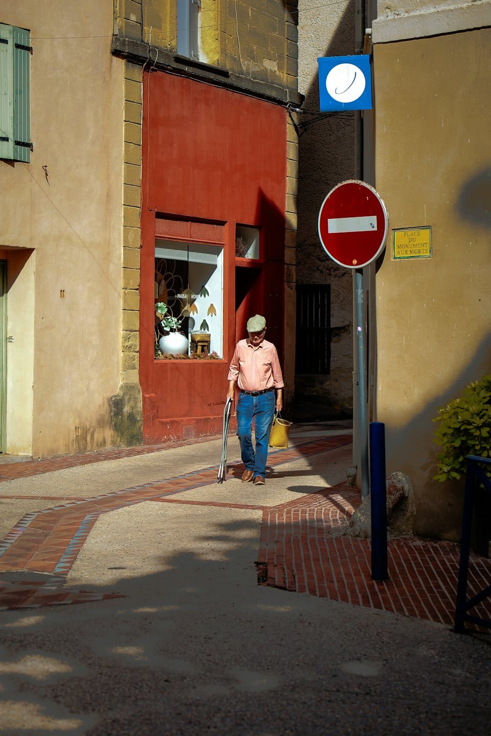 Man walking with a cane and yellow basket down a narrow street.